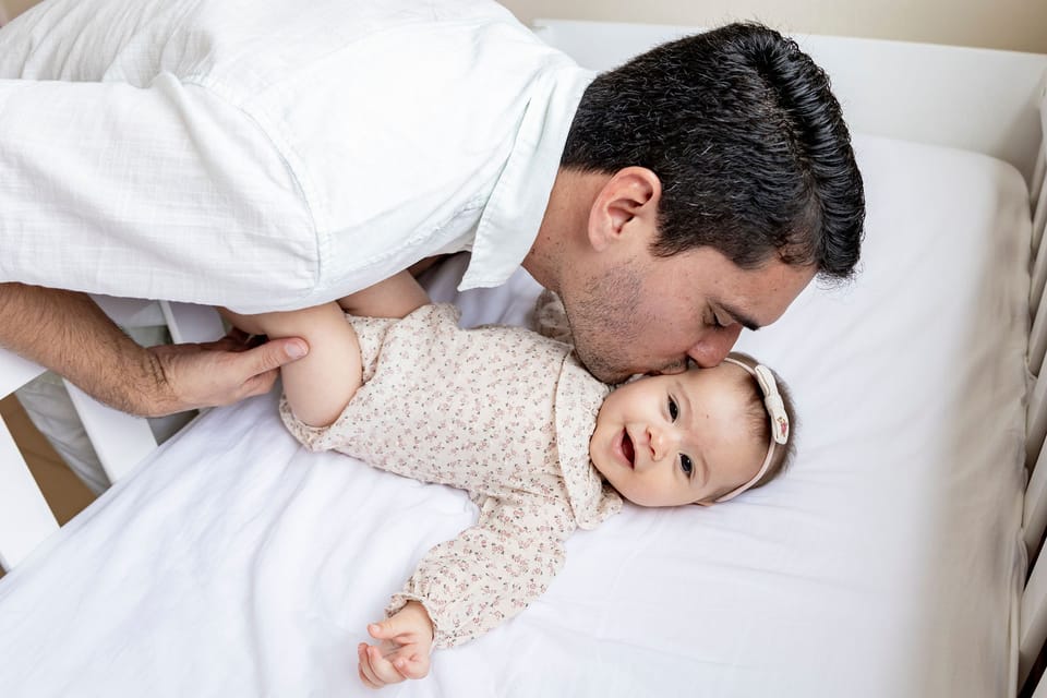 No berço, o pai se debruça e beija a bochecha da filha, que sorri para a câmera.