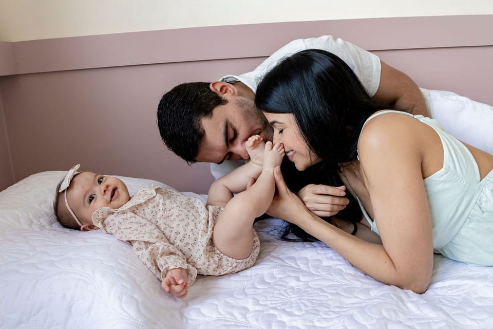 Na cama dos pais, pai e mãe beijam os pezinhos da bebê, que olha para a câmera e sorri.