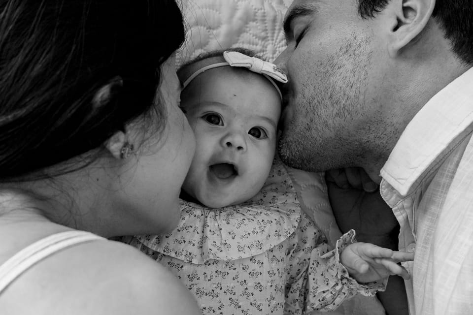 Em preto e branco, pai e mãe beijam as bochechas da bebê, que faz "oh" para a câmera.