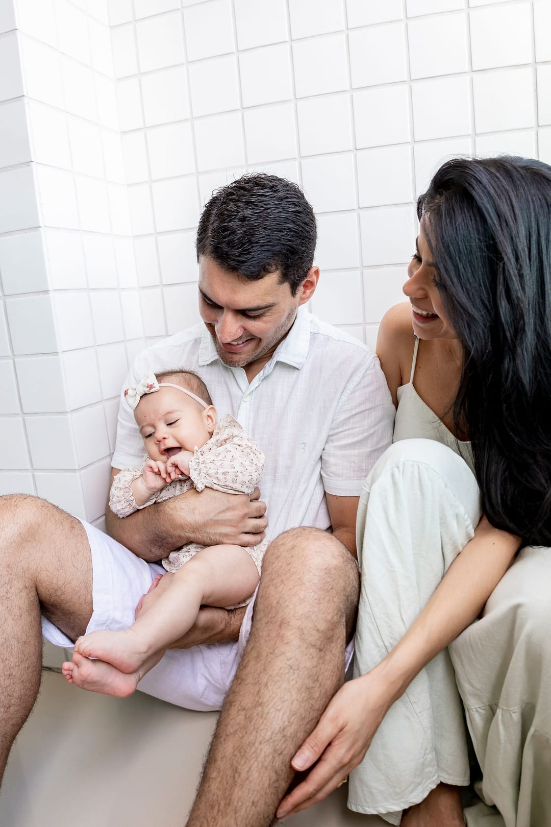Sentados no chão do quarto de azulejos brancos, o pai segura a bebê no colo e a mãe, ao lado, sorri para os dois.
