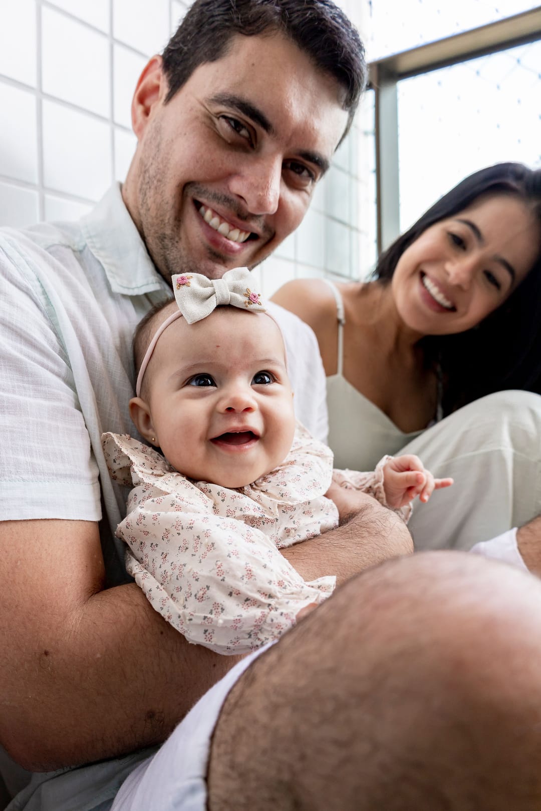 O pai sorri para a câmera com a bebê de laço no colo; a mãe, atrás, ri olhando para ela.