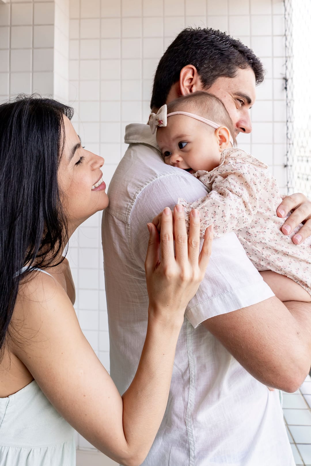 A bebê no ombro do pai, que está de costas; a mãe apoia a mão nas costas dela e sorri.