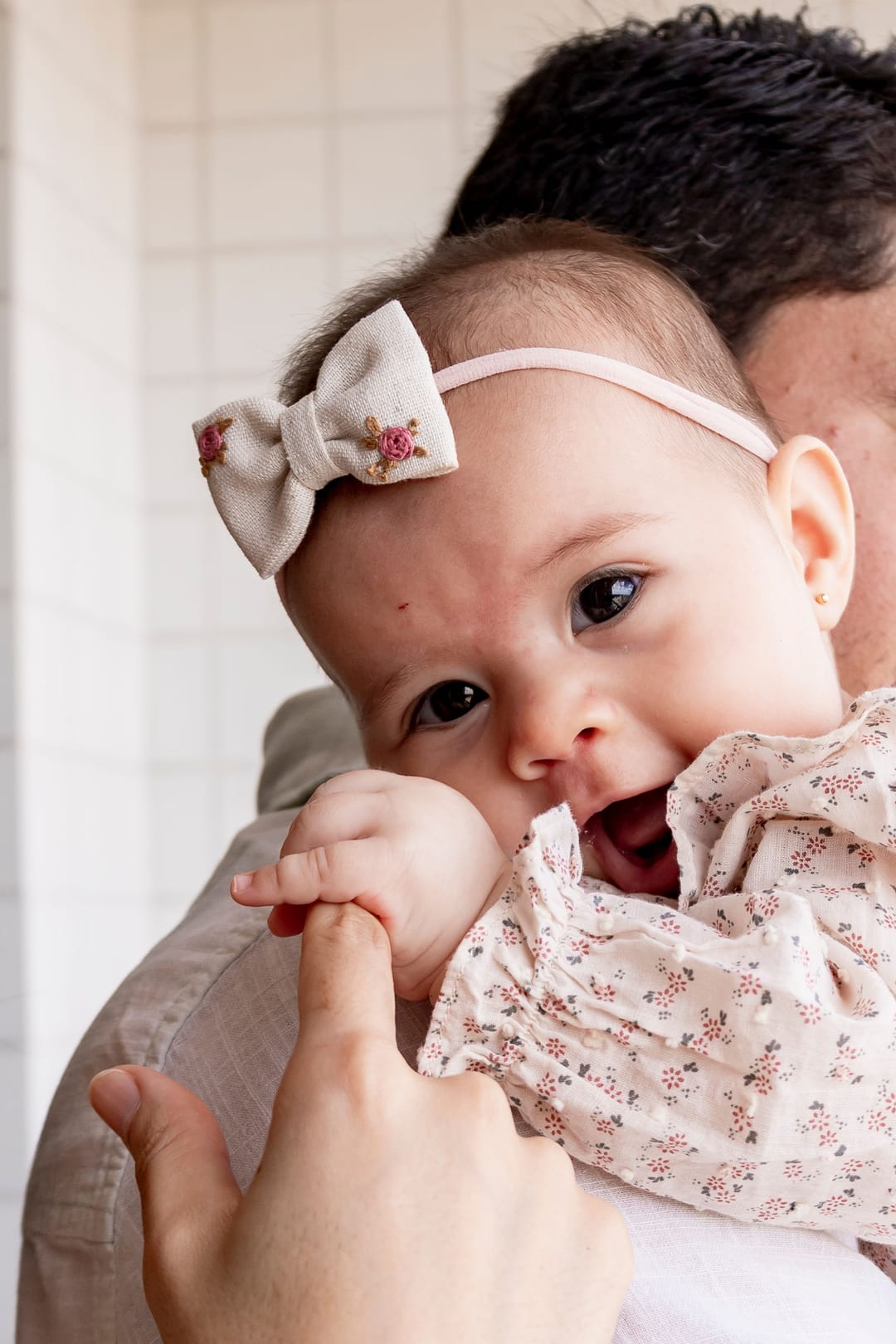 De perto, a bebê de laço segura o dedo do pai e olha para a câmera.