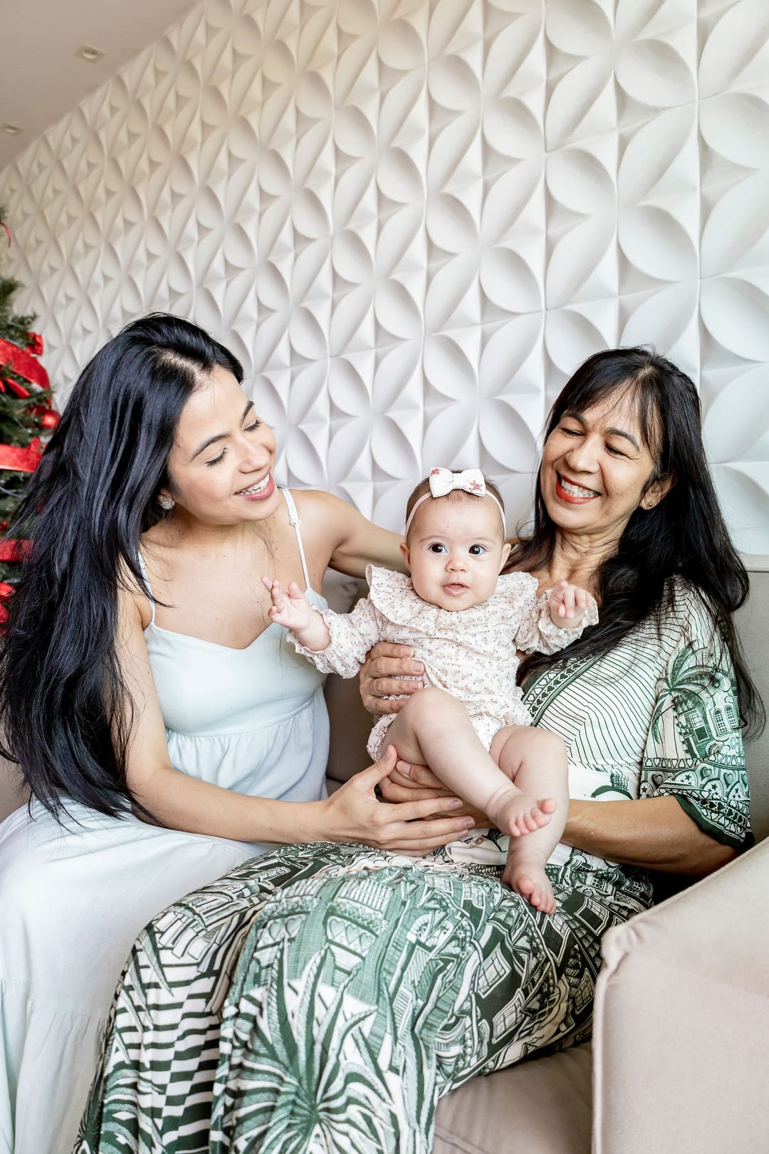 A mãe e a avó, sentadas no sofá diante da parede branca de relevo, seguram a bebê entre elas e sorriem.