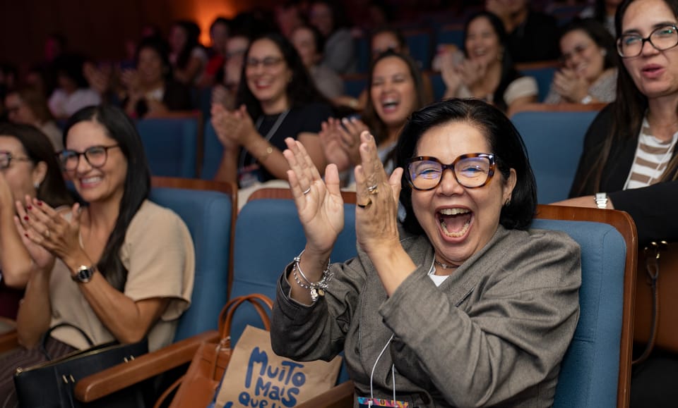 Uma participante de óculos gargalha de boca aberta e aplaude com as mãos no alto.