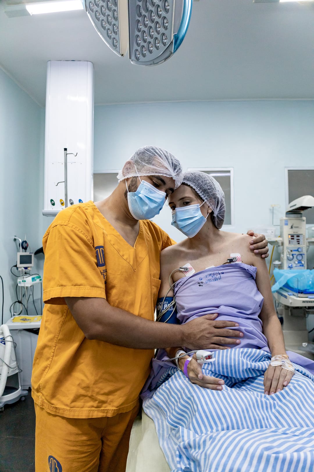 No pré-operatório, ele, de roupa laranja, abraça a mãe na maca; os dois de touca e máscara olham para a câmera.