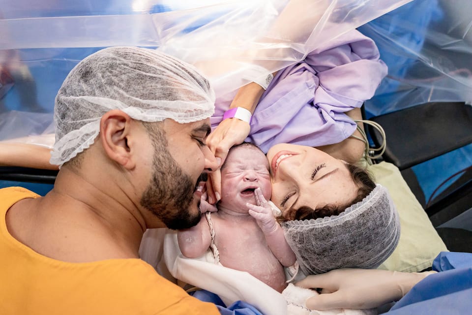 O pai beija o bebê, que chora, e a mãe sorri de olhos fechados.