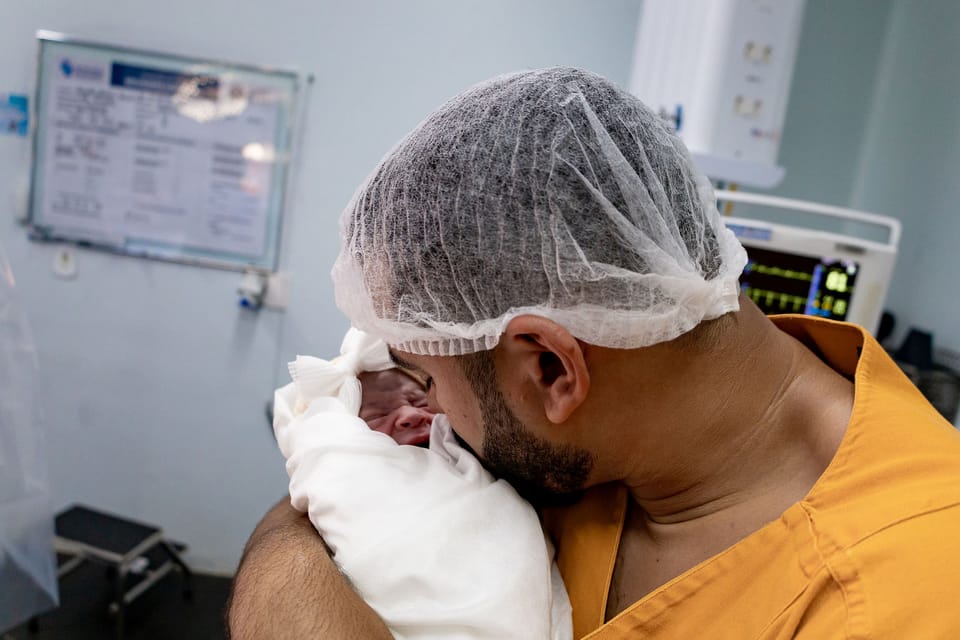 Ele, de touca, segura a recém-nascida enrolada na manta e encosta o rosto nela.