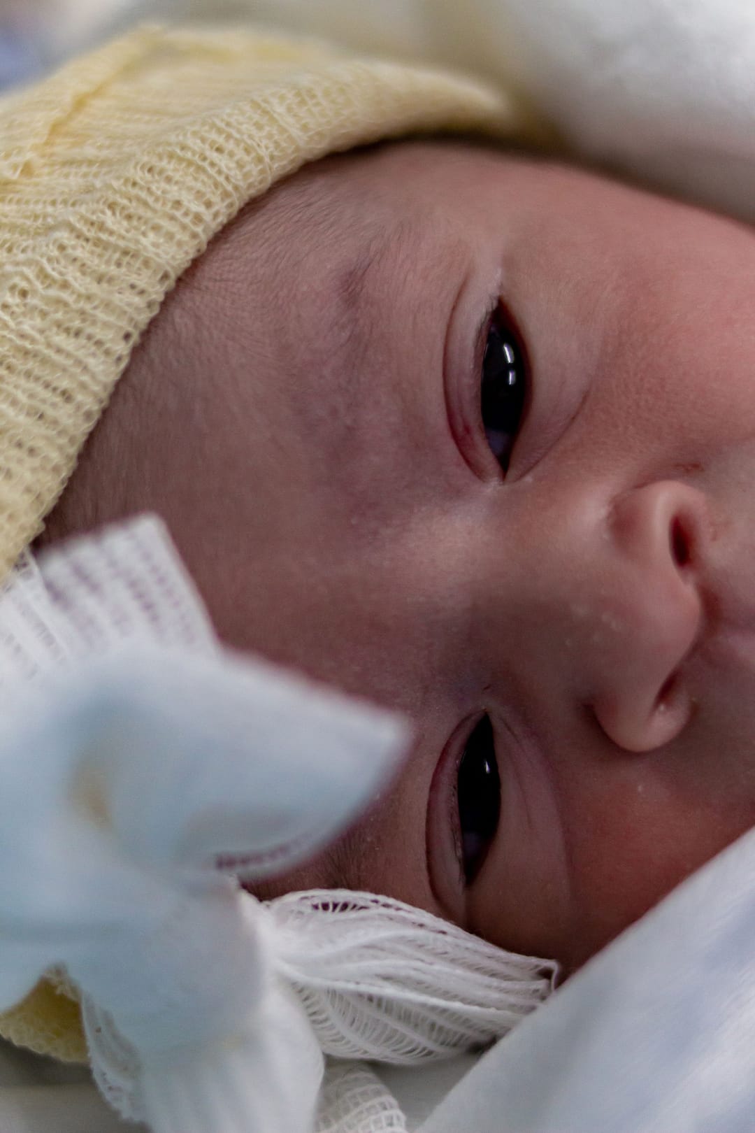 O bebê em close, de olhos abertos, com touca de tricô e um lacinho de gaze.