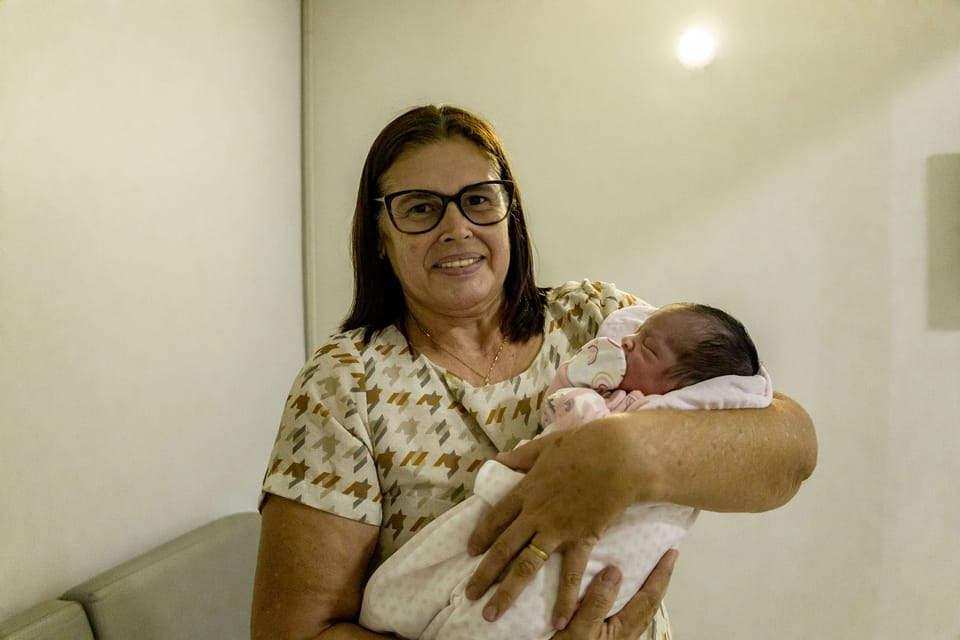 A avó, de óculos, segura a recém-nascida no colo e sorri.