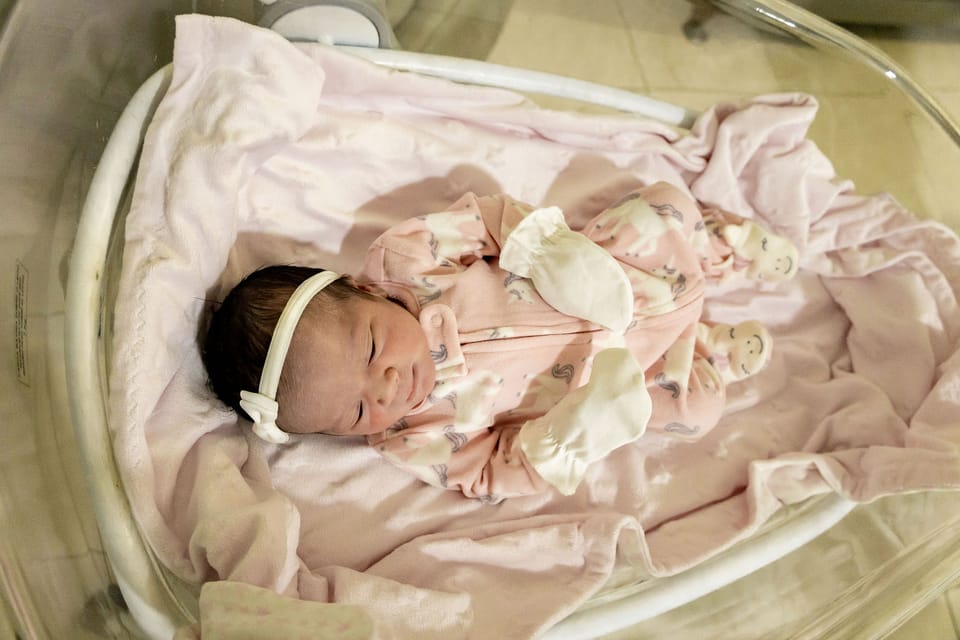 A recém-nascida dorme no berço, vestida de rosa, com uma faixa de laço na cabeça.