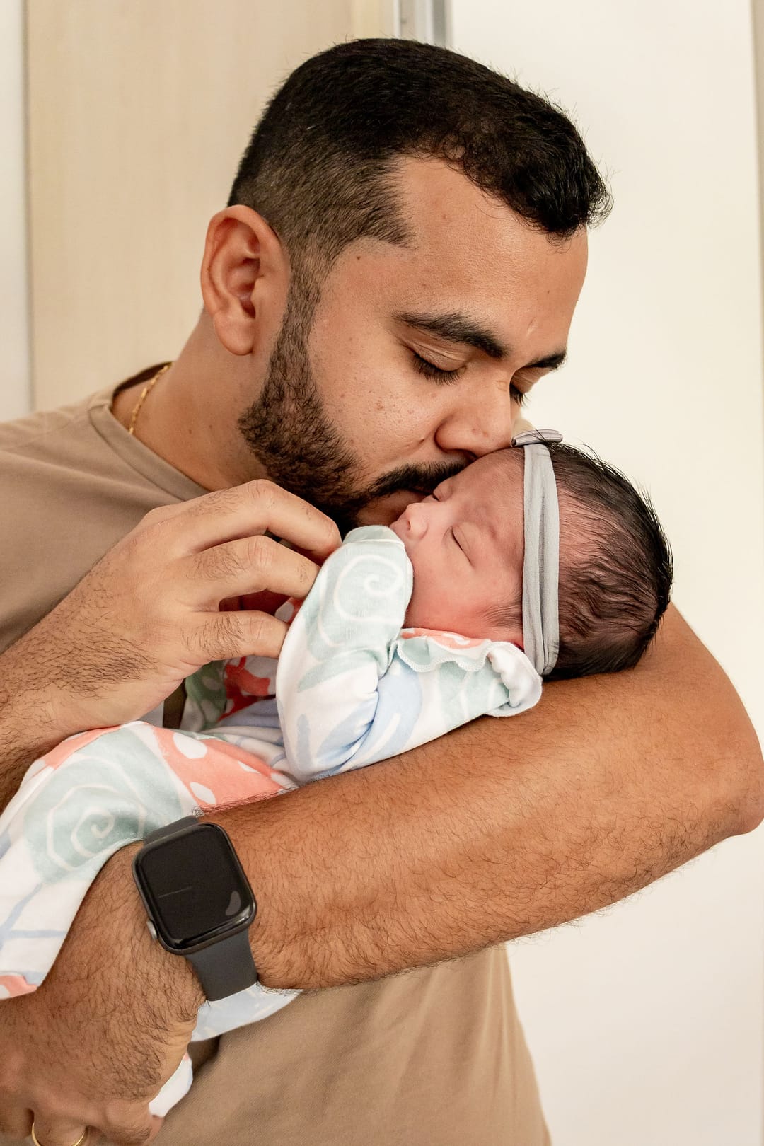 O pai, de olhos fechados, beija a cabeça do bebê aninhado no peito.