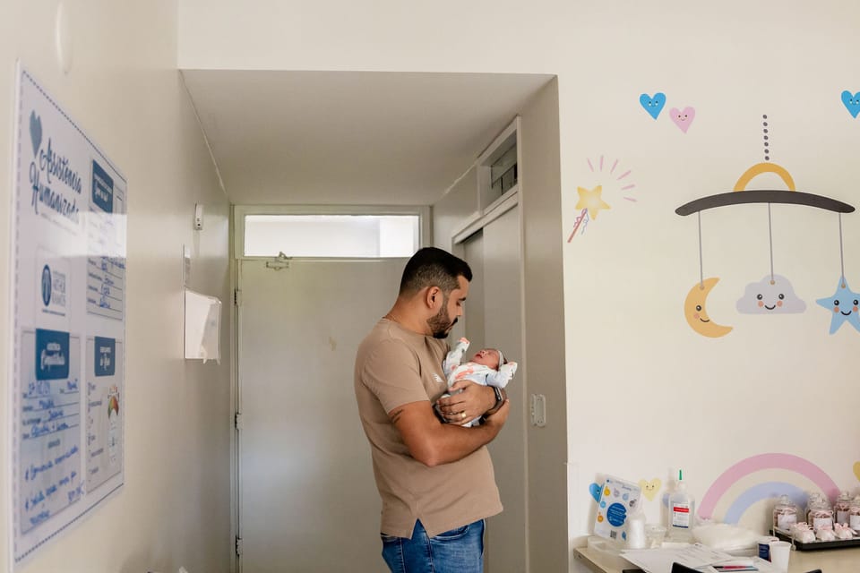 No quarto decorado com nuvens e arco-íris, o pai segura a recém-nascida junto à porta.