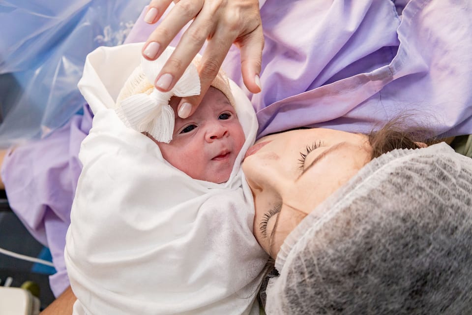 O bebê de olhos abertos para a câmera, com o rosto colado ao da mãe, que ajeita o lacinho.