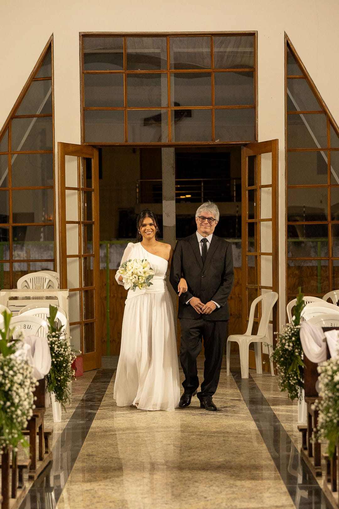 A noiva e o pai atravessam o portal de madeira e vidro da igreja; ela sorri.