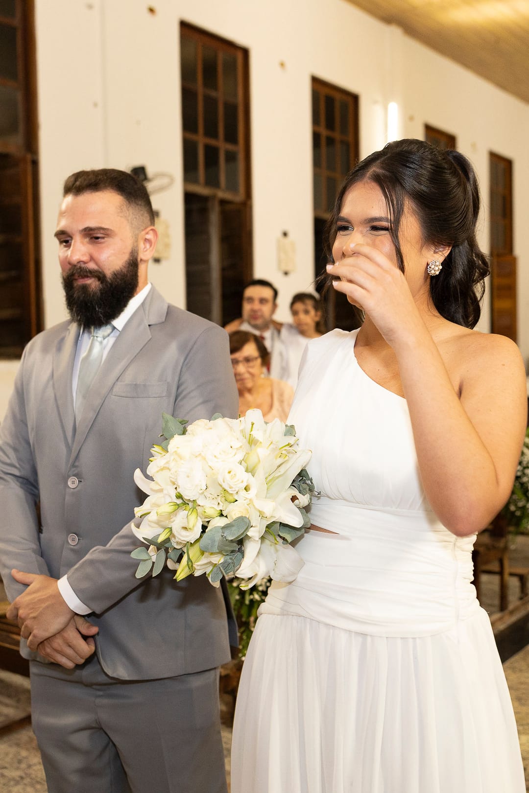 A noiva enxuga uma lágrima com o buquê de lírios na mão; o noivo, emocionado, ao lado.