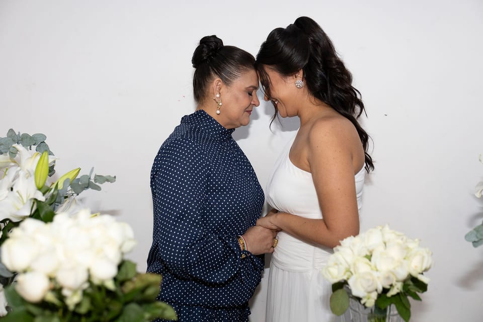 A mãe e a noiva, de mãos dadas, encostam testa com testa entre rosas e lírios desfocados.
