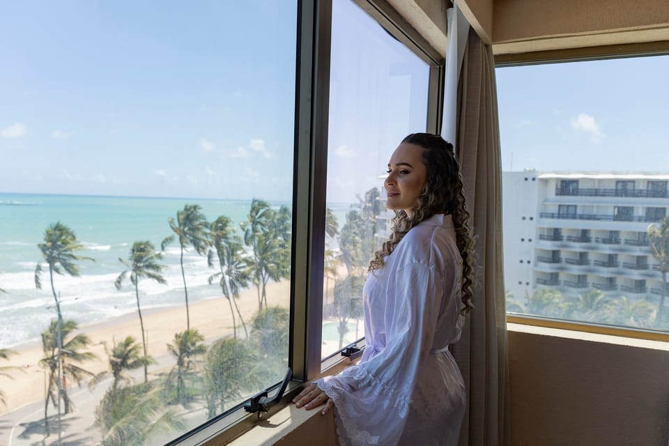 De robe, a noiva olha pela janela de canto do hotel a orla com coqueiros e o mar verde.