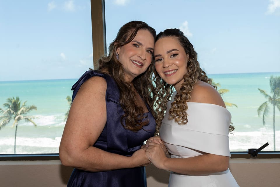 Mãe e filha, a noiva, sorriem juntas diante da janela com o mar e os coqueiros.