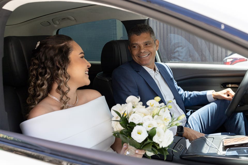 A noiva sorri no banco do carona; o pai, ao volante, sorri para ela.