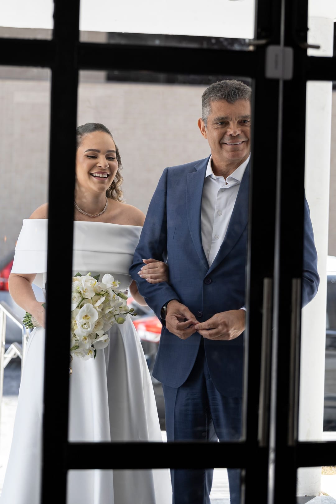A noiva de braço com o pai, vistos pela porta de vidro de caixilho preto.