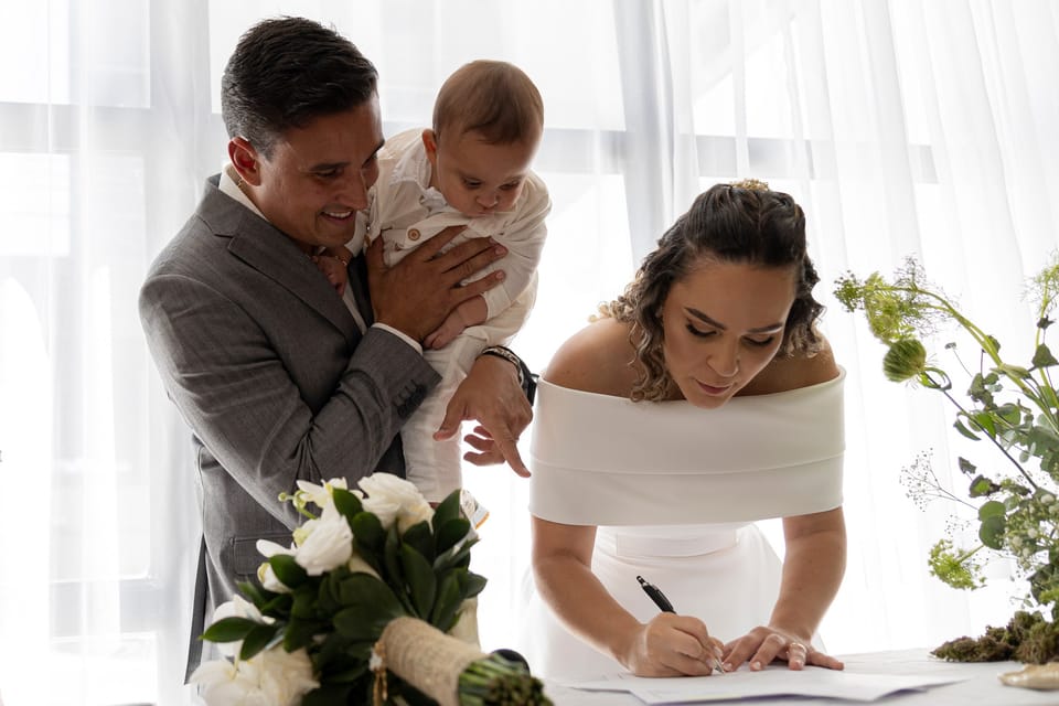 A noiva assina; o noivo, com o bebê no colo, sorri e aponta o papel.