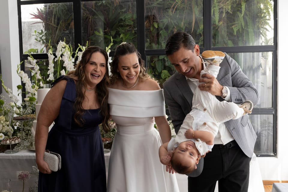A noiva, a mãe dela e o noivo riem juntos; ele segura o bebê de ponta-cabeça.