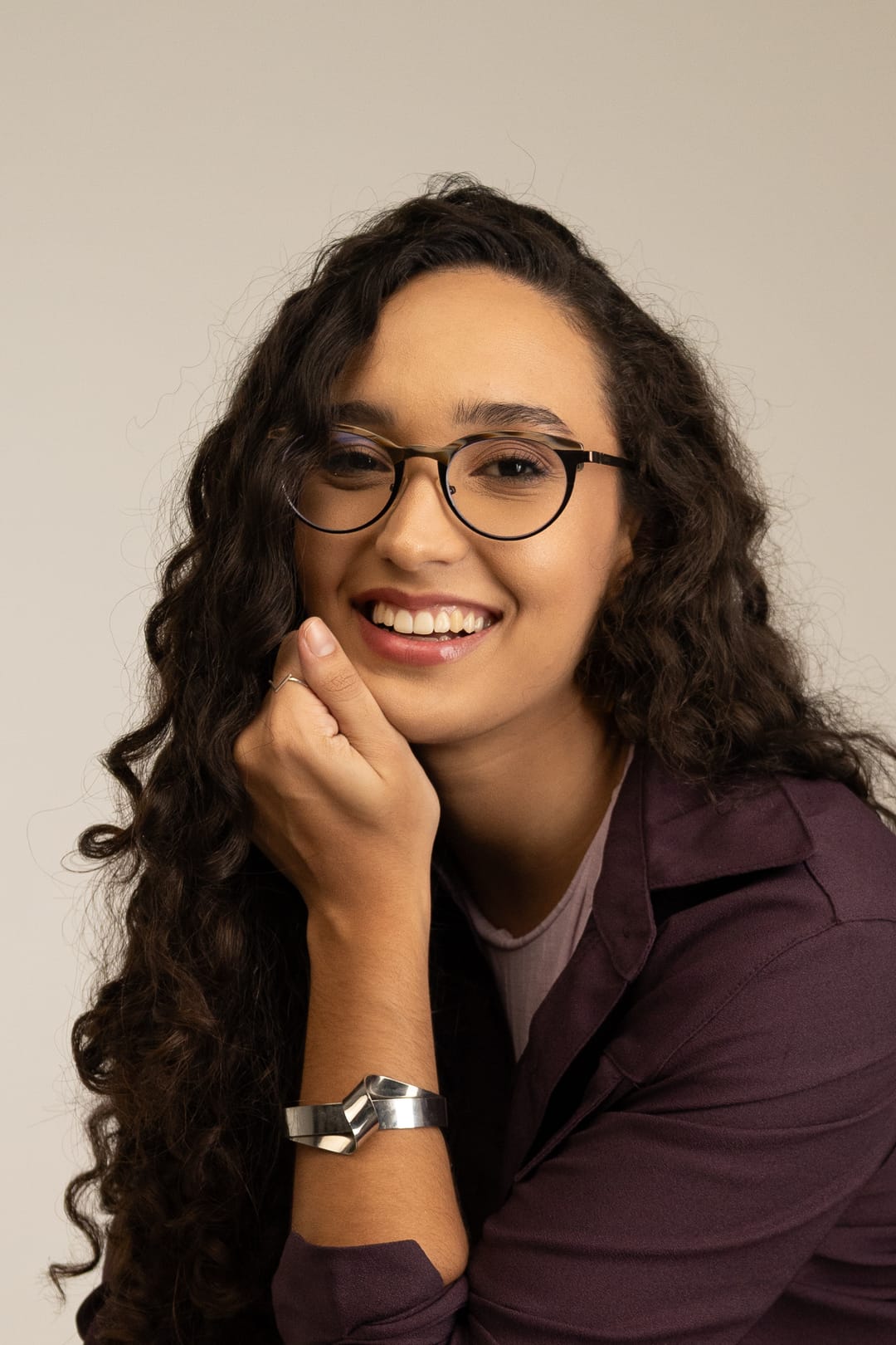 A Marcela sorri de óculos, com a mão no queixo e o cabelo cacheado solto.