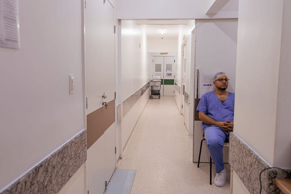 O pai, de pijama cirúrgico, sentado sozinho no fim de um corredor.
