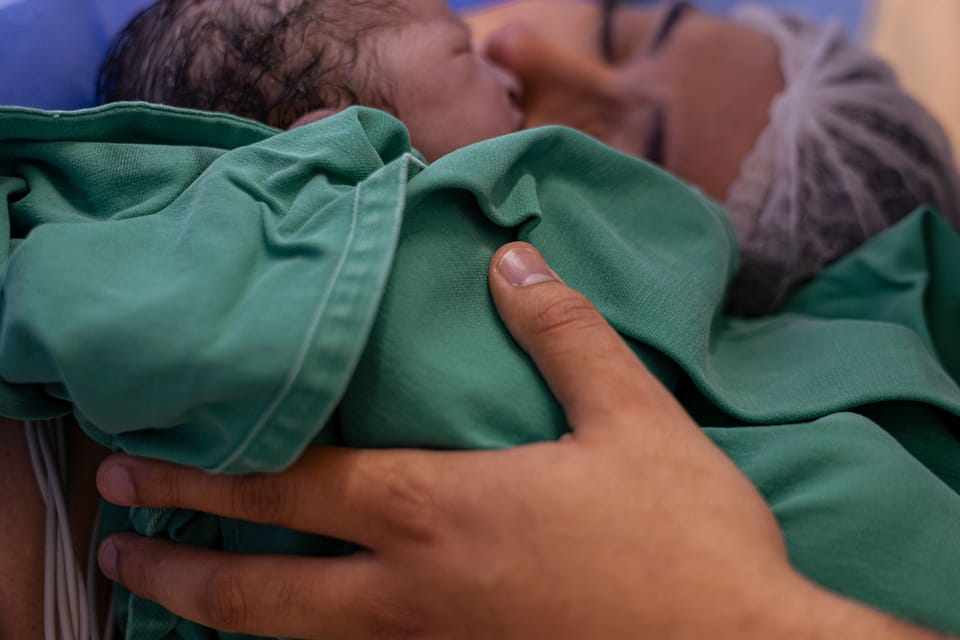 Enrolado no pano verde, o bebê encosta no rosto da mãe; a mão do pai o segura por cima.