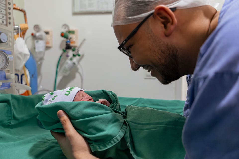 O pai, de touca e óculos, se debruça sorrindo sobre o bebê enrolado no pano verde.
