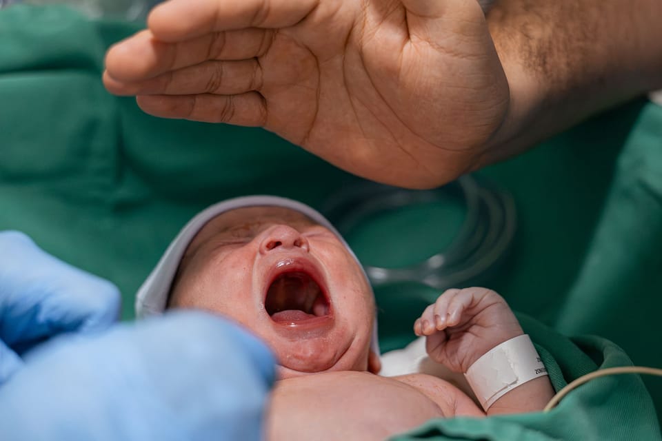 O bebê chora de boca aberta sob uma mão estendida, no berço aquecido.