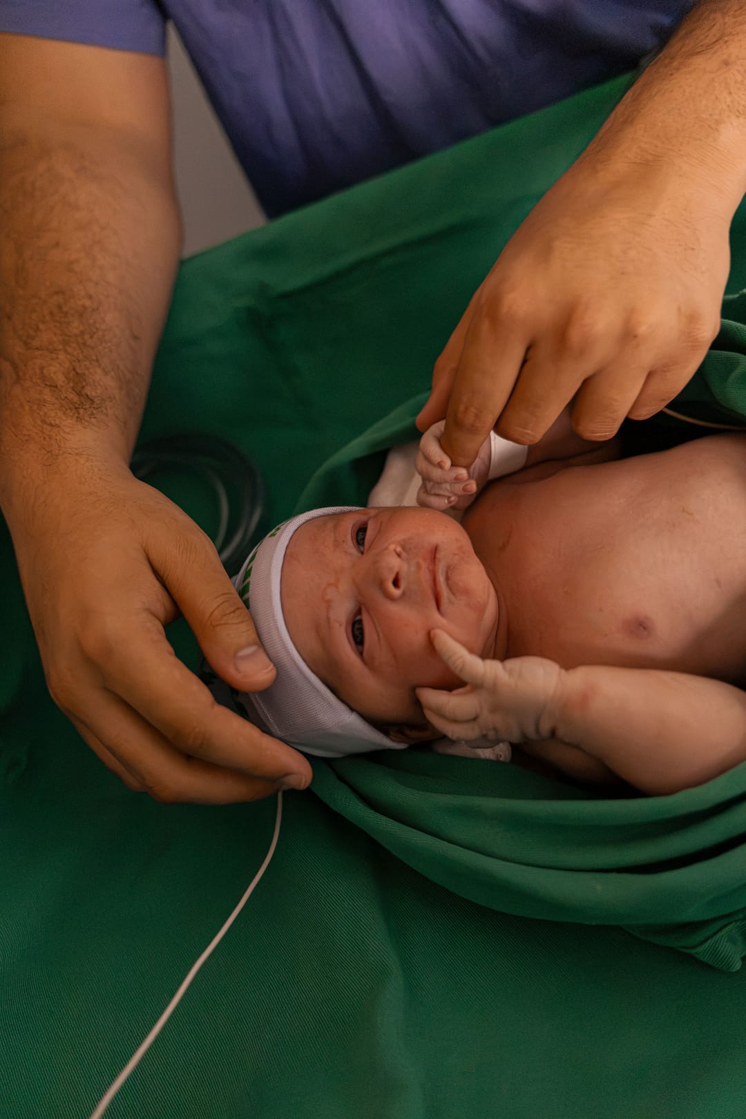 No berço aquecido, o bebê de olhos abertos segura o dedo do pai, que emoldura a cabeça dele com as mãos.