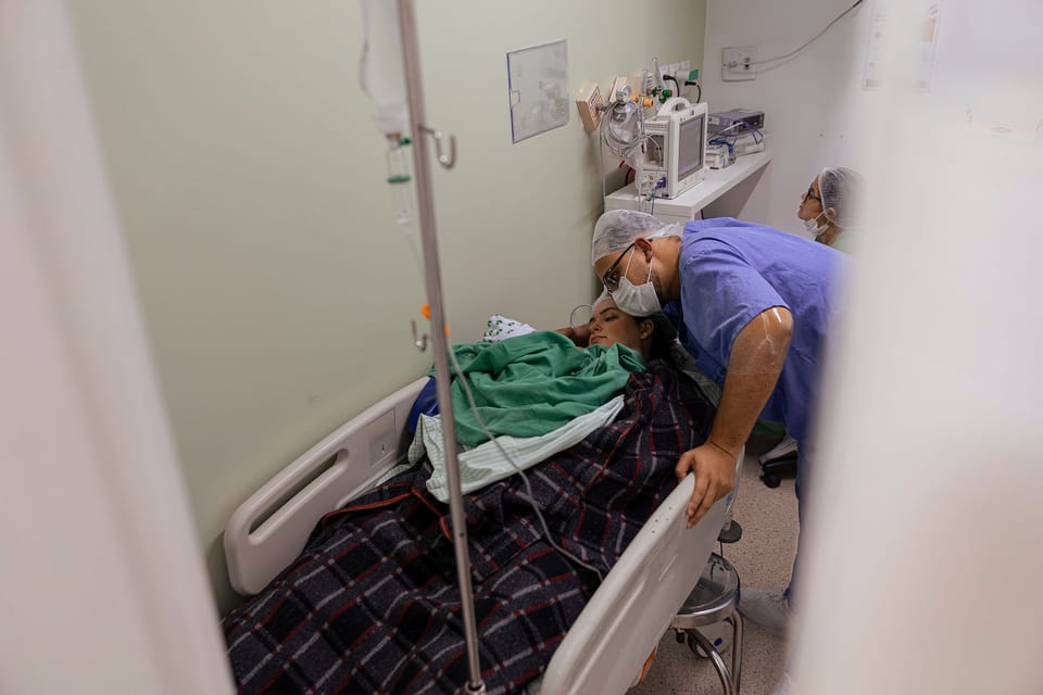Pelo vão da cortina, o pai se debruça sobre a mãe e o bebê na maca da recuperação.