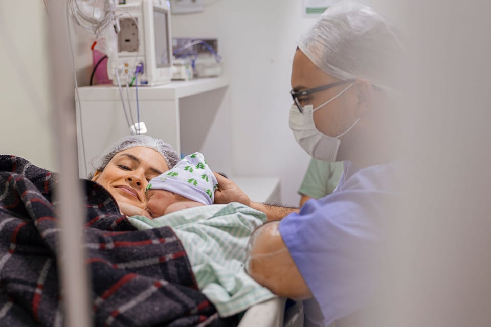 Pela cortina, a mãe sorri com o bebê de touca no peito, e o pai se aproxima.