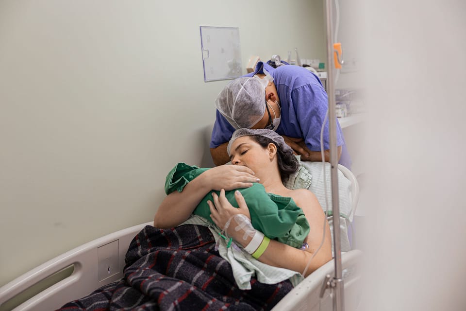 O pai se debruça sobre a mãe, que abraça o bebê na maca.