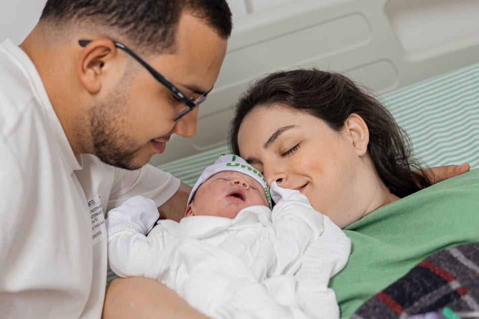 Os três na cama: ele sorri olhando o filho, e ela encosta o rosto de olhos fechados.