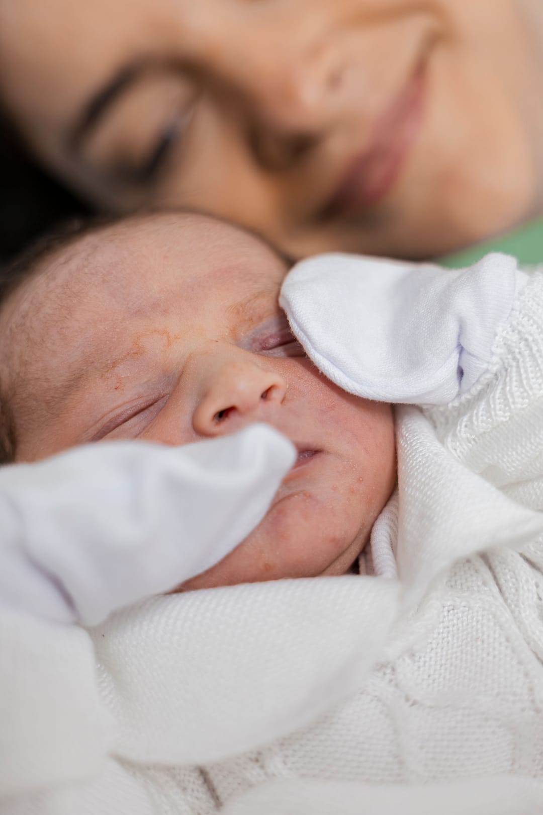 O bebê nítido, de luvinha, e o sorriso da mãe desfocado no alto.