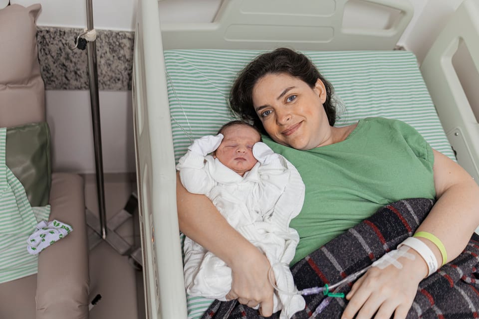 Na cama do quarto, a mãe sorri deitada ao lado do recém-nascido.
