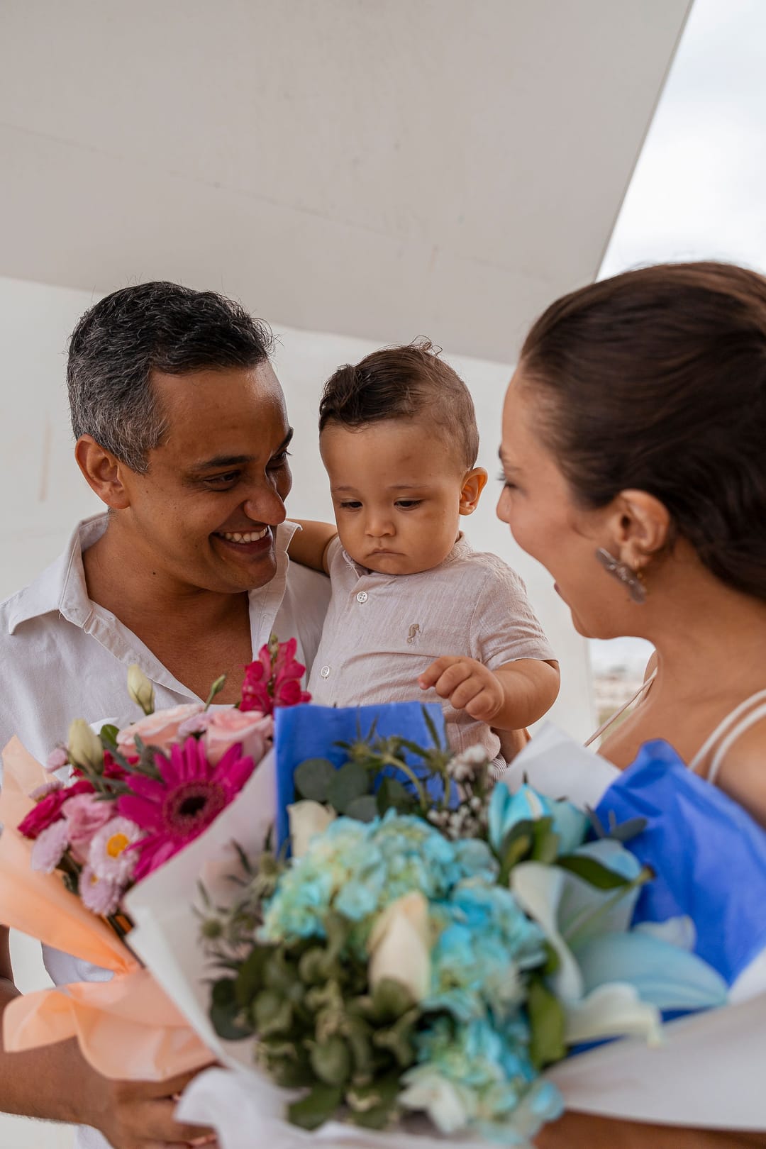 O pai, com o filho no colo, e a mãe sorriem entre os dois buquês, um azul e um rosa.