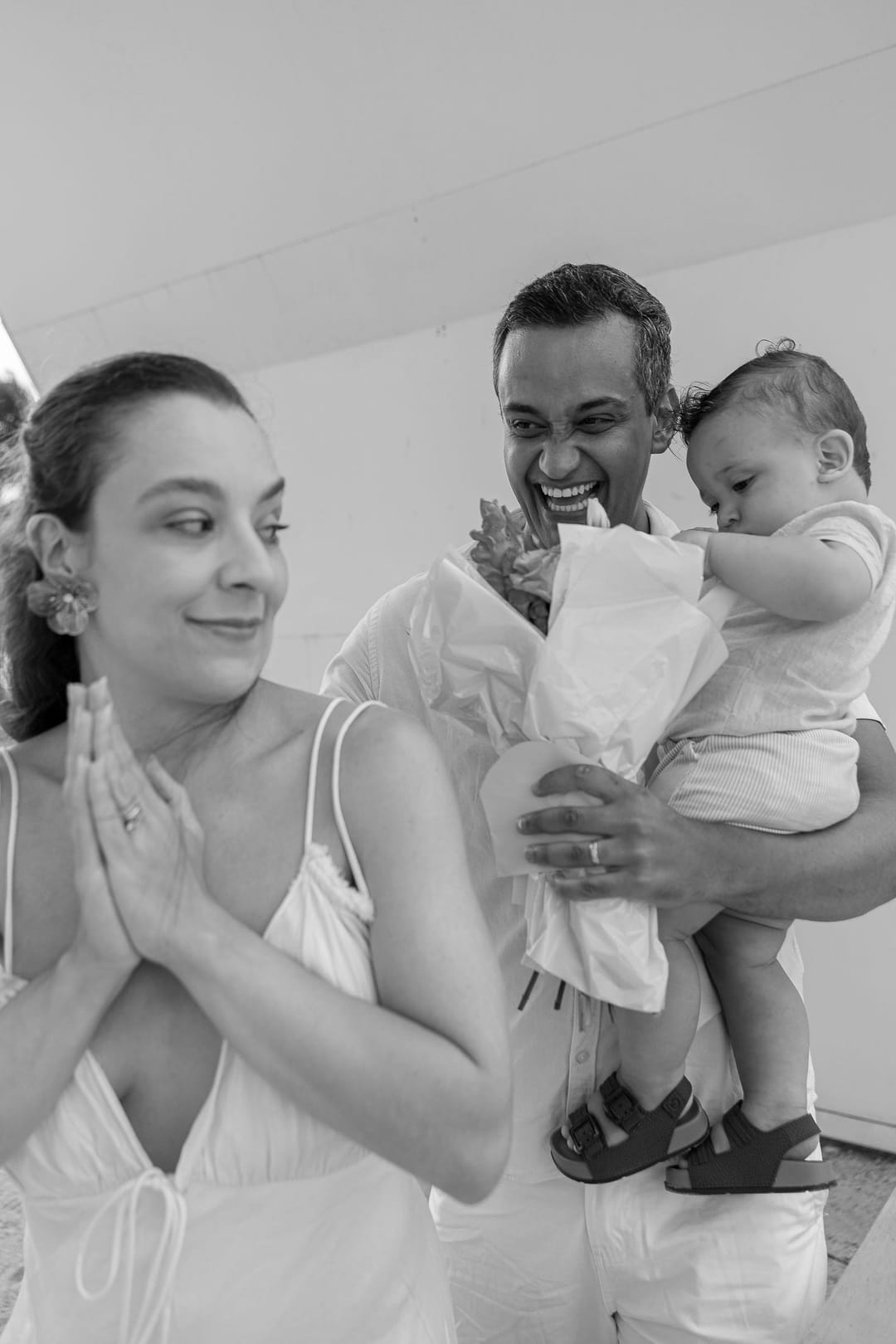 A mãe junta as mãos diante do rosto, ansiosa; ao lado, o pai segura o filho e o buquê rosa.