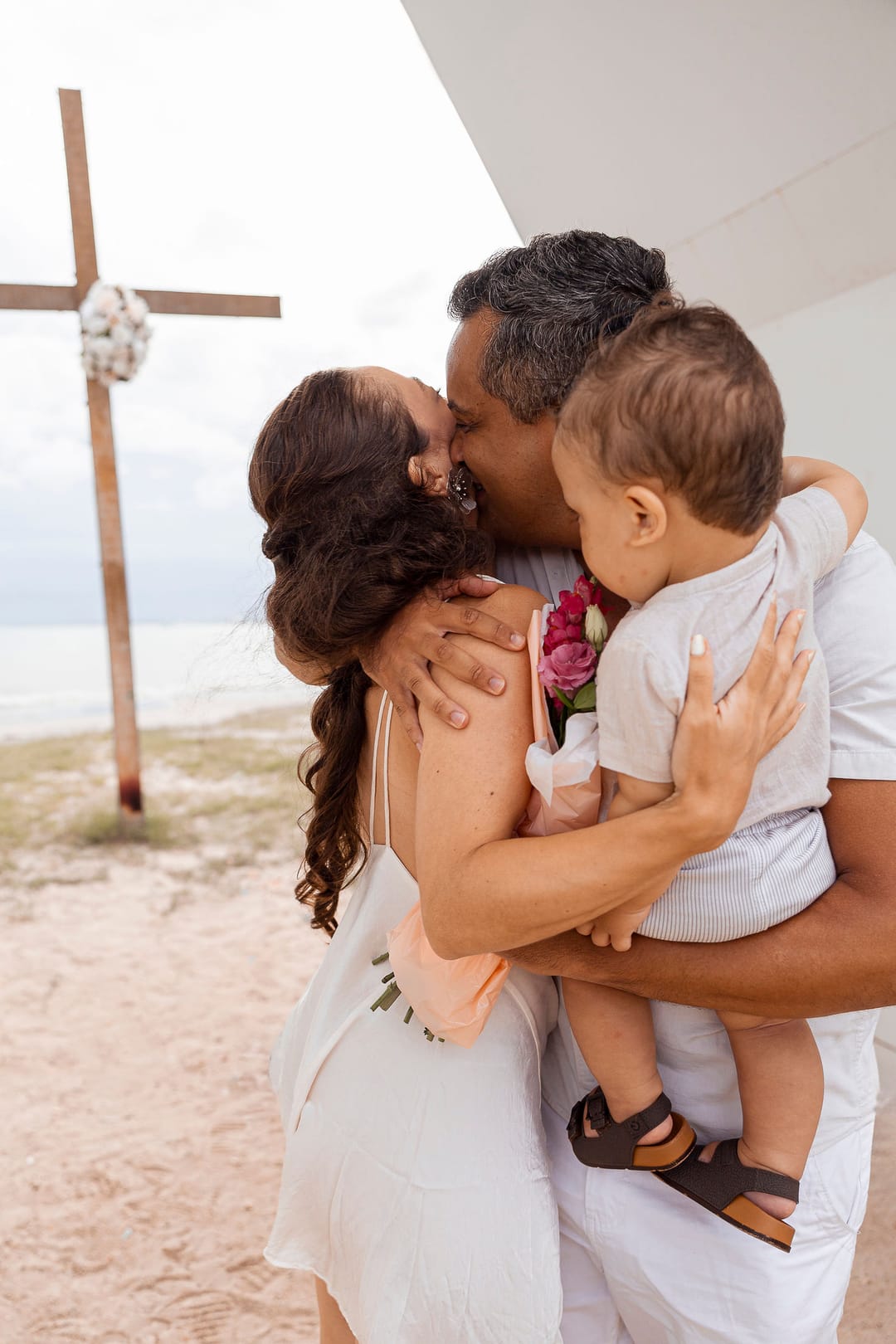 Na capela, a mãe abraça o pai e o filho, com o buquê rosa entre eles.