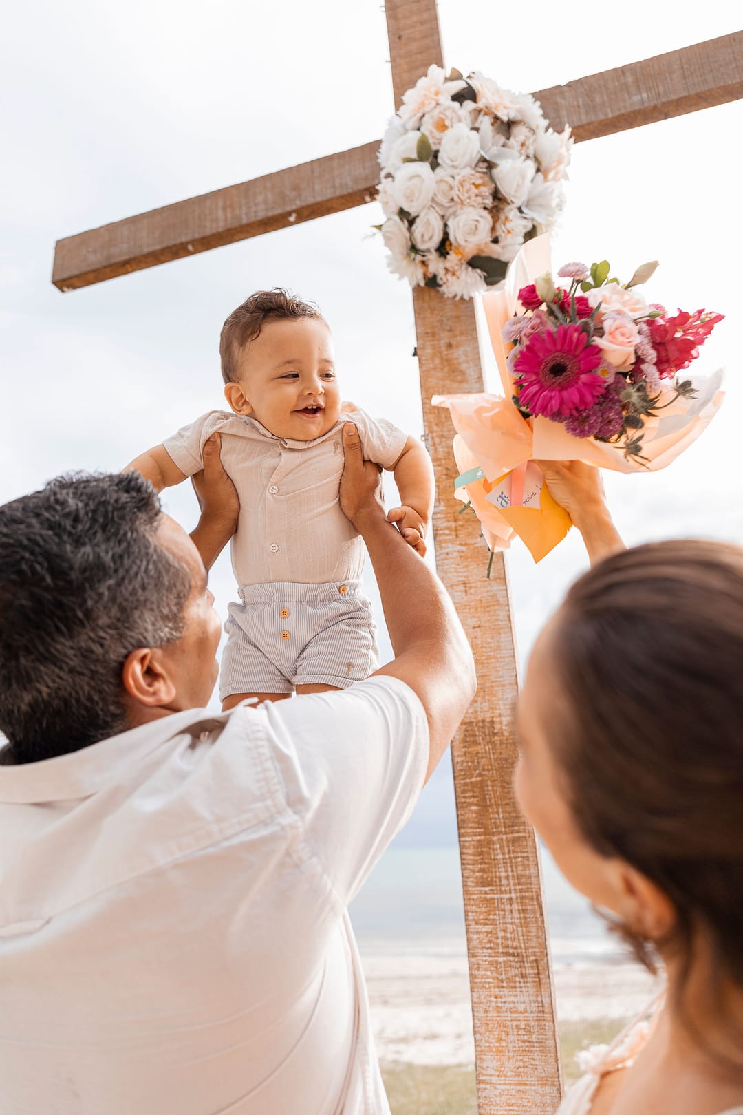 O pai ergue o filho no alto diante da cruz enfeitada de flores; a mãe olha de baixo.