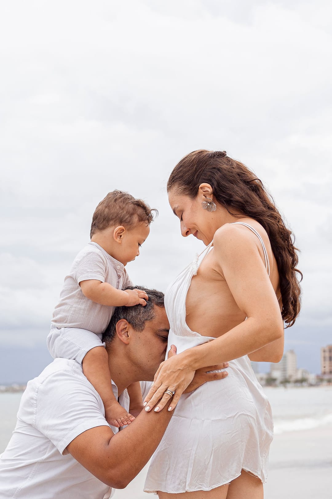Na orla, o pai beija a barriga; o filho, nos ombros dele, olha para baixo.