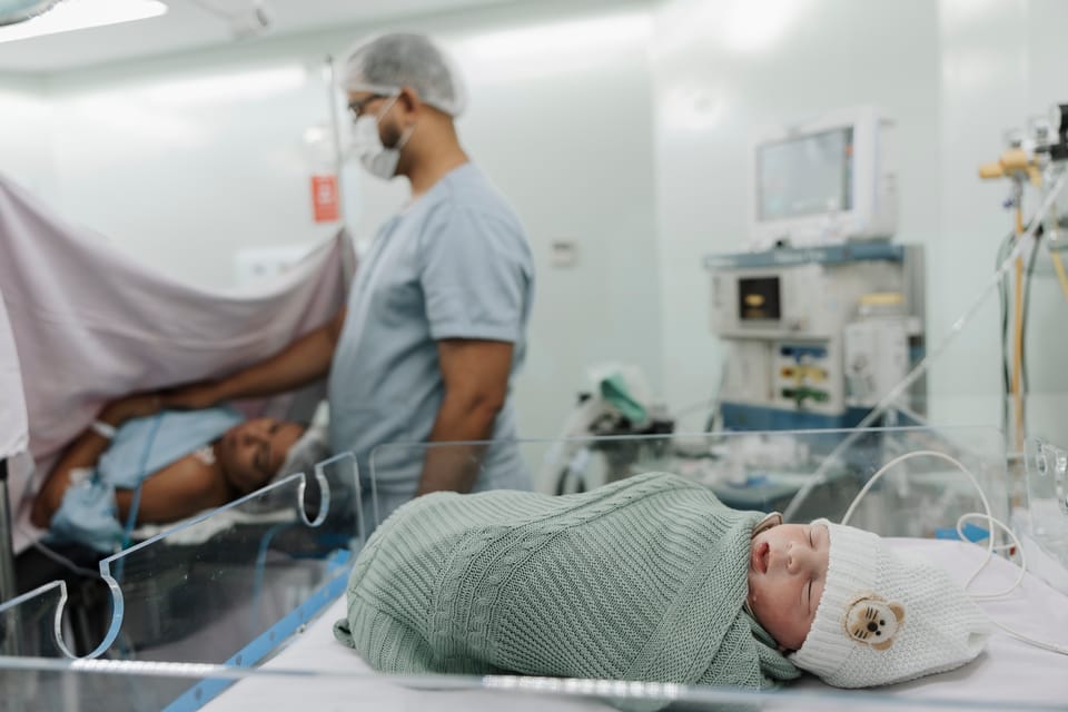O bebê no berço de acrílico, em primeiro plano; ao fundo, o pai ao lado da mãe.