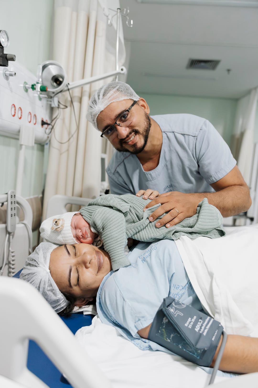 O pai sorri ao lado da mãe, que segura o bebê deitada na maca.