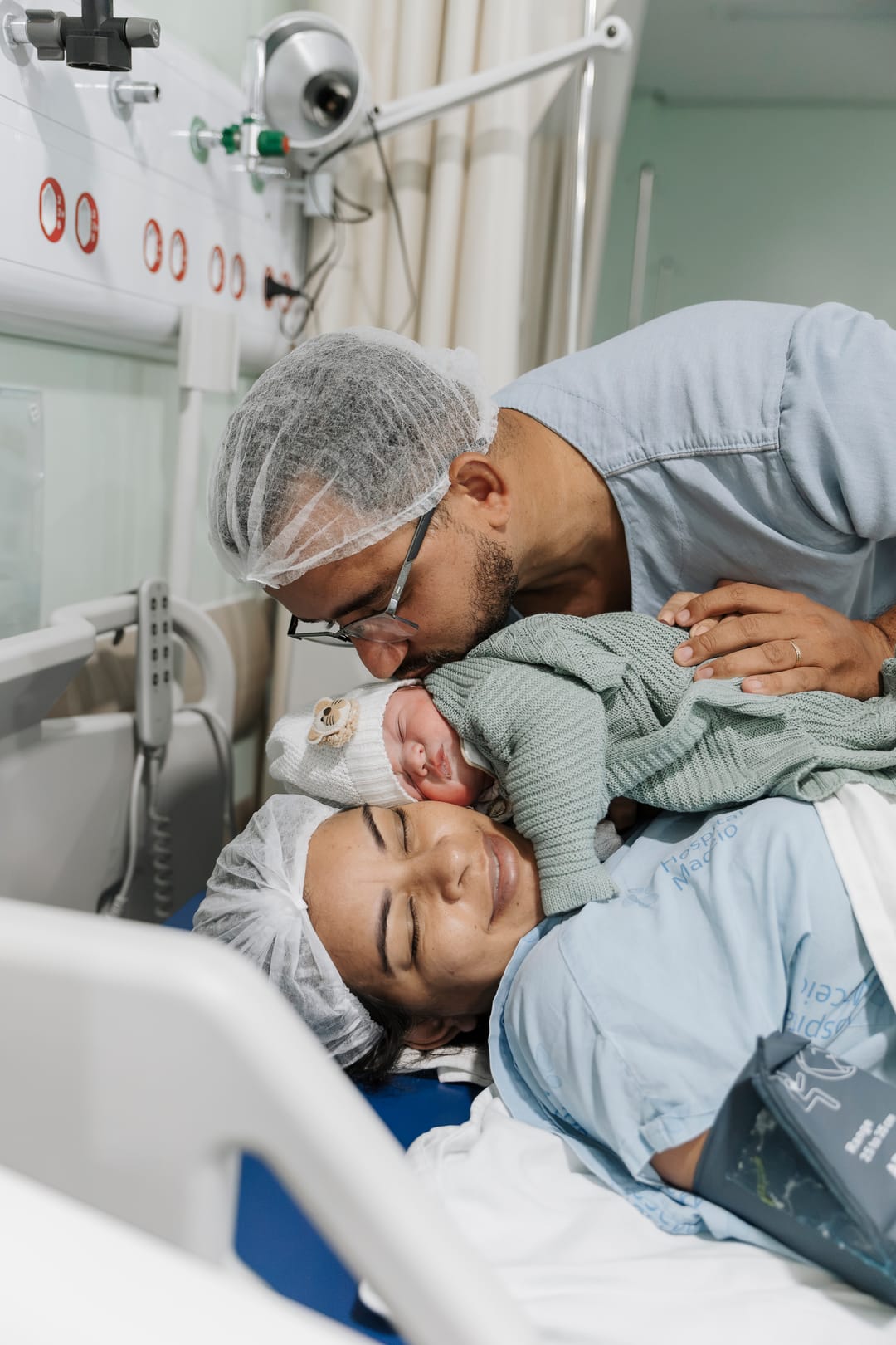 O pai beija a cabeça do bebê, deitado no ombro da mãe, que sorri de olhos fechados.