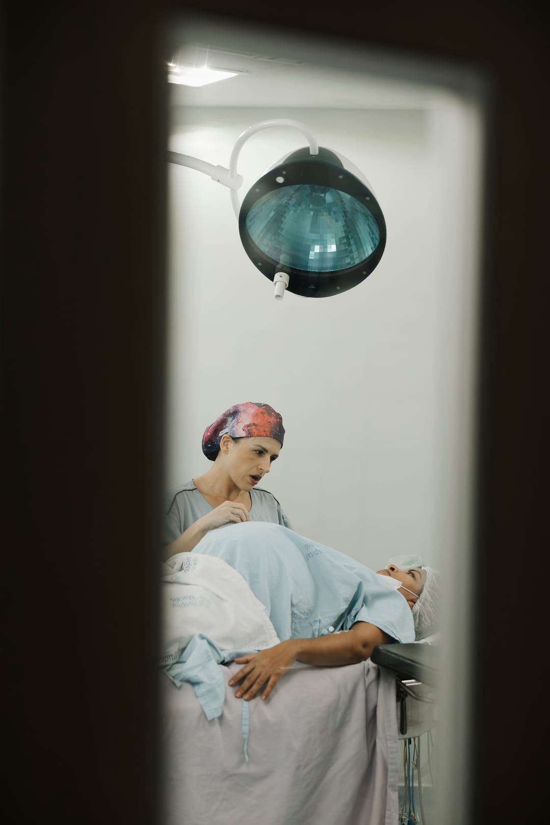 Pela janela estreita da porta, a mãe deitada no centro cirúrgico, sob o foco, com uma profissional ao lado.