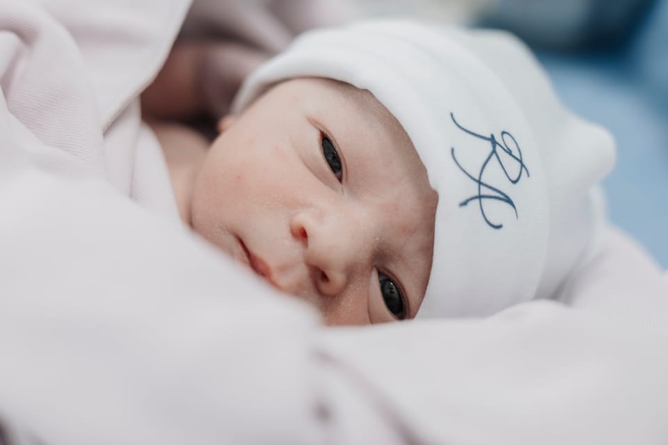 Enrolado na manta rosa, de touca bordada, o bebê abre os dois olhos para a câmera.