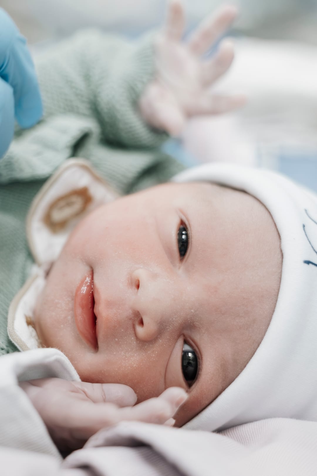De tricô verde-sálvia e touca de ursinho, o bebê olha para a câmera com a mãozinha no ar.
